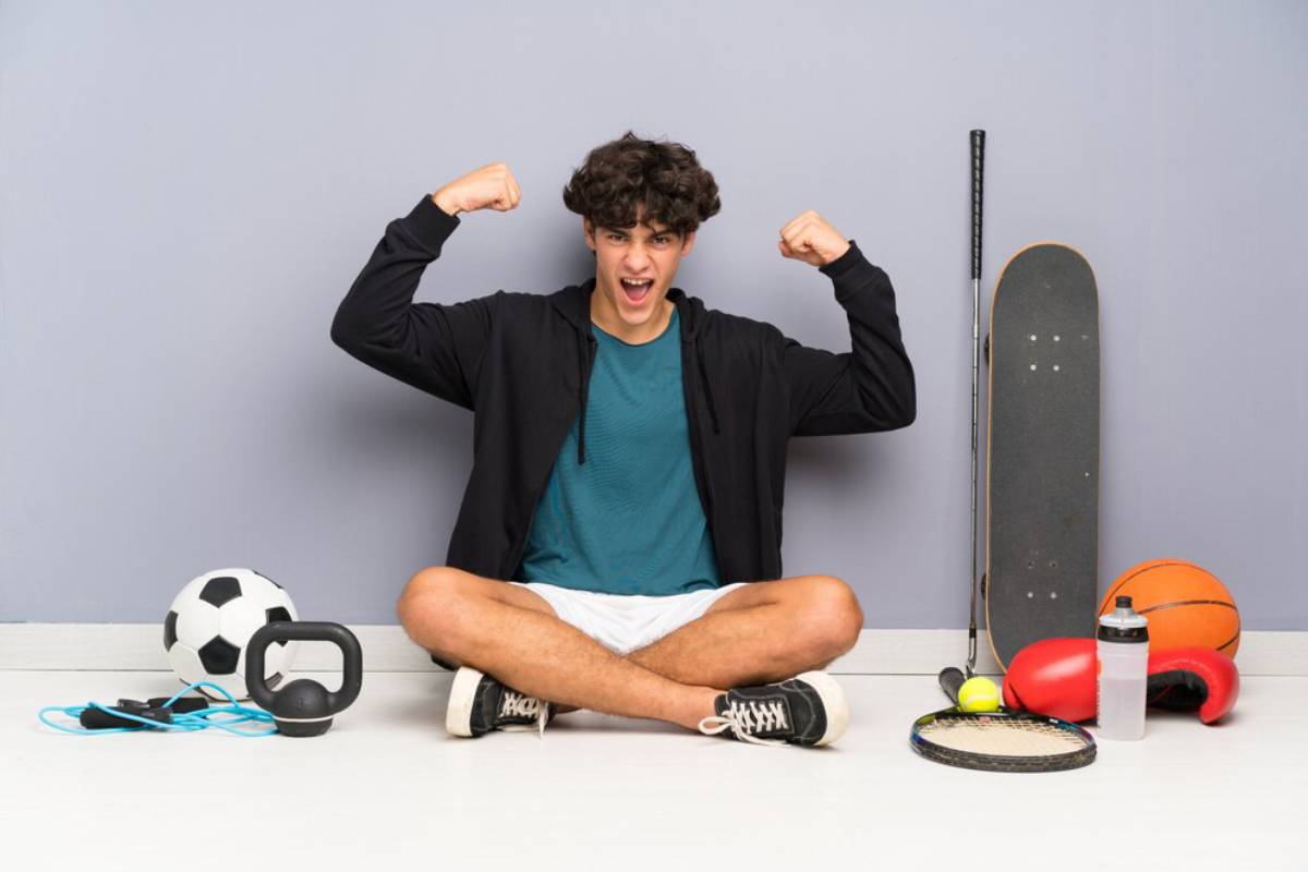 Young man raising fists in victory, sitting with sports equipment like a basketball, soccer ball, skateboard, and racket against a gray background.